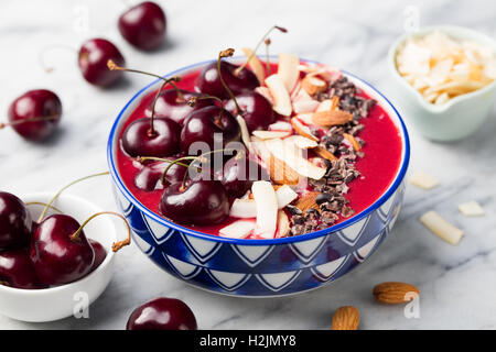 Smoothie-Schüssel mit frischen schwarzen Kirschen, Kokosflocken, Mandeln und Kakao Nibs. Marmor-Hintergrund Stockfoto