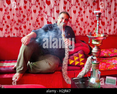 Ein irakischer Mann sitzt auf einer roten Couch Rauchen ist Shisha, Wasserpfeife, in einem Café in Kuala Lumpur, Malaysia. Stockfoto