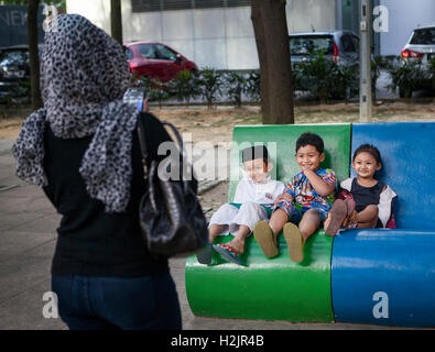 Happy muslimische Kinder sitzen auf einer Parkbank, während ihre Mutter ihr Bild in Kuala Lumpur, Malaysia. Stockfoto