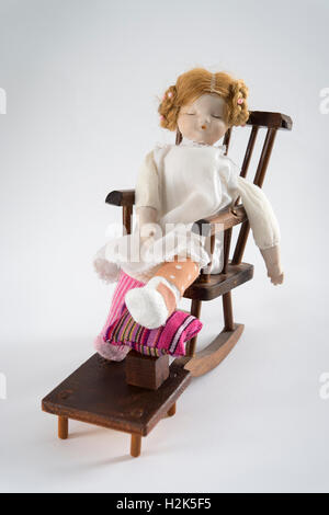 Puppe schlafend im Schaukelstuhl, bandagierte Bein auf Hocker mit Kissen, weißer Hintergrund Stockfoto