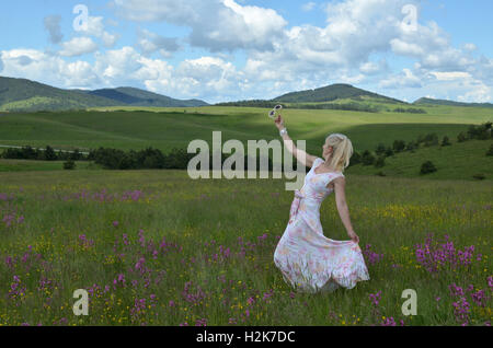 Lady in einem Kleid mit floralem Muster tanzt auf der Wiese, mit der Sonnenbrille in einer hand Stockfoto