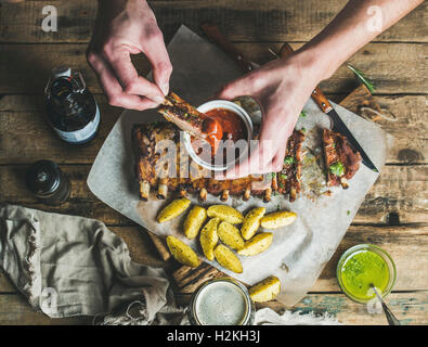 Menschen essen gebratene Schweinerippchen mit Kartoffeln, Knoblauch, Rosmarin und grüner Kräutersauce auf rustikalen Holztisch. Mann ' s Hände Dipp Stockfoto