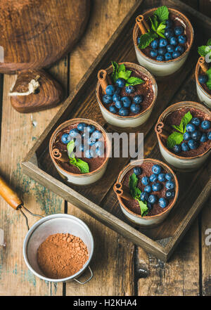 Hausgemachte Tiramisu Dessert in Gläser mit Zimt, Minze und frische Heidelbeeren in Holztablett und Sieb mit Kakaopulver über r Stockfoto