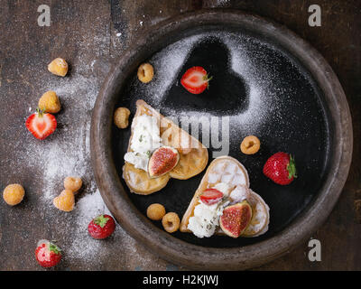 Wafer als Herzform mit gelbe Himbeeren, Erdbeeren, in Scheiben geschnittene Feigen, Ricotta-Käse und Zucker Pulver auf Ton Fach über dunkel Stockfoto