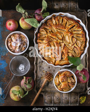 Hausgemachte große und kleine Kuchen Apfelkuchen in weißer Keramik Formen auf Vintage Metalltablett mit frischen Äpfeln mit Blättern, Puderzucker ein Stockfoto