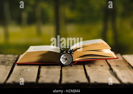 Dickes Buch liegen offen auf hölzernen Oberfläche, alte altmodische Nachttisch Uhr sitzt daneben Magiekonzept schießen Stockfoto