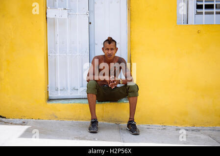 Junger Mann mit Tattoo sitzt vor der Haustür des gelben Hauses in Santiago De Cuba, Kuba. Stockfoto