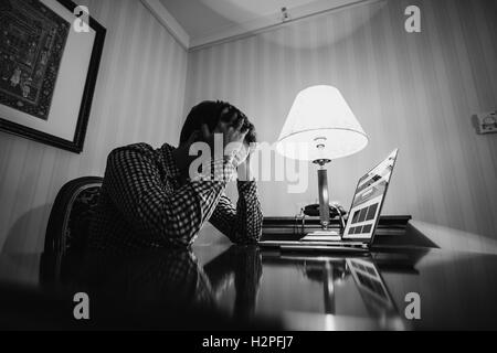 Erschöpft. Frustrierter junge Mann mit Händen auf dem Kopf und Augen zu halten beim Sitzen am Arbeitsplatz geschlossen. Schwarz weiß Foto. Stockfoto