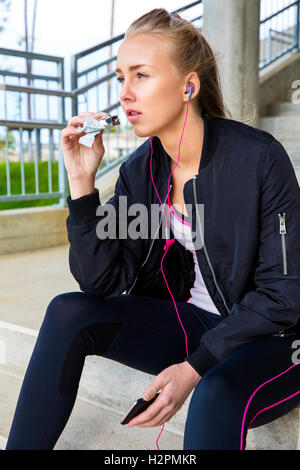 Sportliche Frau Essen Protein Bar beim hören Musik auf Schritte Stockfoto