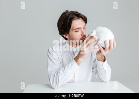 Junge Brünette Mann küssen gefälschte Schädel auf den grauen Hintergrund isoliert Stockfoto