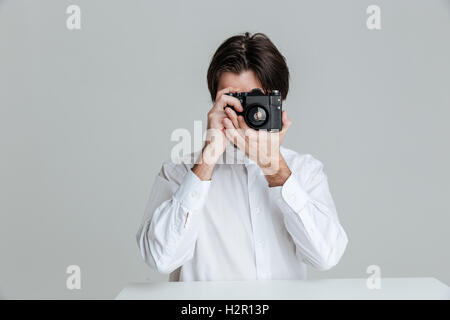 Hübscher Junge Brünette Mann am Tisch sitzen und machen Fotos mit der Kamera auf den grauen Hintergrund isoliert Stockfoto
