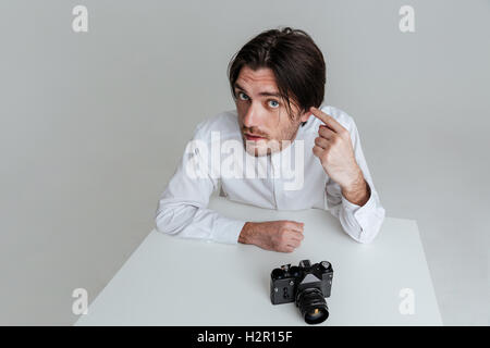 Junge Borsten Brünette Mann zeigte auf sein Ohr, während sitzen am Tisch auf dem grauen Hintergrund isoliert Stockfoto