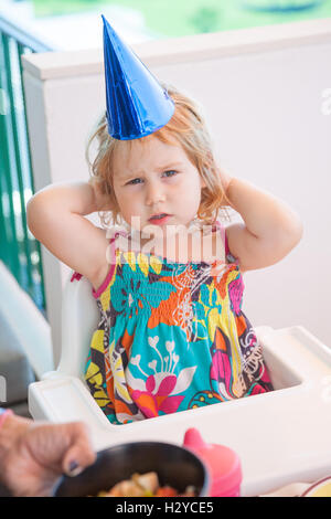 zwei Jahre alte blonde Kind mit hellen blauen Karton Partyhut sitzen in weißen Hochstuhl ernsthaft Stockfoto