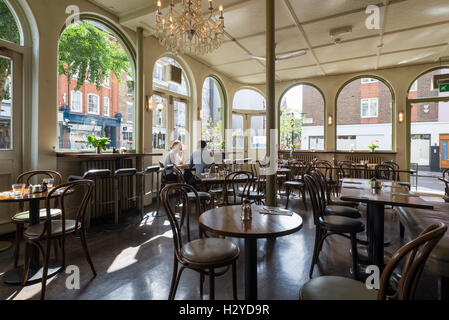 Junges Paar sitzt auf Holzstühlen am runden Tisch zum Frühstück im stilvollen Coco Momo Cafe in Marylebone High Street, Westminster, London, UK Stockfoto