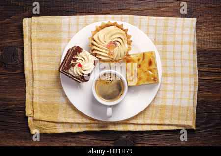 Drei Kuchen und Kaffeetasse im weißen Teller auf einer gelben Tischdecke. Ansicht von oben Stockfoto