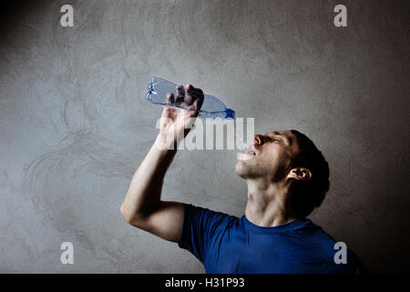 Muskel-Mann trinkt Wasser aus Plastikflasche auf graue Wand Hintergrund Stockfoto