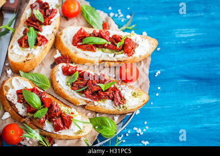 Bruschetta mit Frischkäse, Pesto, frischem Basilikum und getrockneten Tomaten auf blauen rustikalen Holztisch Stockfoto