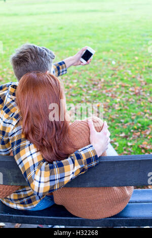 Rückansicht eines jungen Paares, wobei ein Selbstporträt auf einem Smartphone. Sie sind auf einer Bank in einem Park kuscheln. Stockfoto