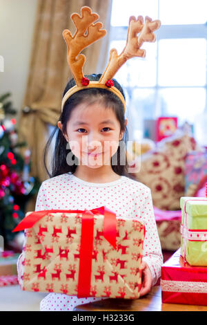 Niedlichen chinesischen Mädchen festliche Geweihe tragen. Sie ist ein Geschenk hochhalten und mit einem frechen Lächeln auf ihrem Gesicht in die Kamera schaut Stockfoto