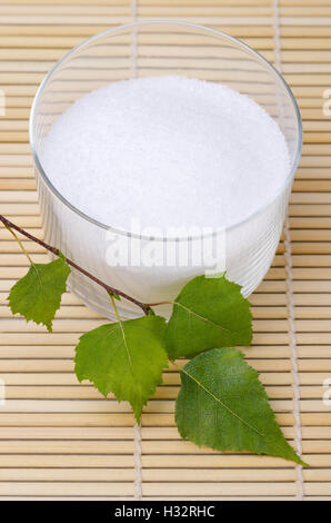Xylitol Birkenzucker in eine Glasschüssel mit Birke Blätter auf einer Bambusmatte. Weißer granulierter Zucker Alkohol, Süßstoff. Stockfoto