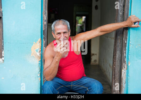 Kubanische Mann trägt eine rote Weste, eine Zigarette rauchend sitzt in seiner Tür Trinidad Kuba Stockfoto