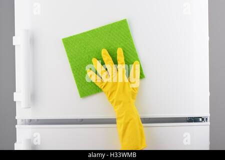 Zarte Frauenhand in schützenden gelben Latex Handschuh Reinigung weißer Kühlschrank mit grünes Tuch auf grauem Hintergrund Stockfoto