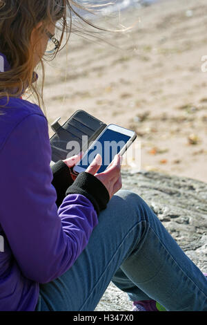 Frau sitzt auf dem Felsen und mit ihrem Smartphone. Wind weht ihr Haar. Stockfoto