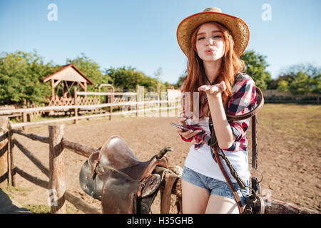 Hübsche Rothaarige Cowgirl in Strohhut senden Luft Kuss auf dem Zaun sitzend Stockfoto