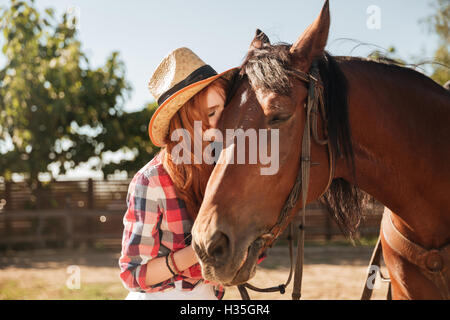 Schöne rothaarige junge Frau Cowgirl stehen und ihr Pferd küssen Stockfoto