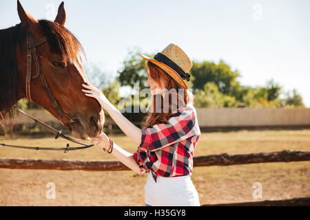 Lächelnd rothaarige junge Frau Cowgirl kümmert sich um ihr Pferd auf Bauernhof Stockfoto