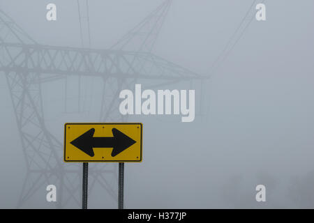 Gelbe Richtungsanzeiger in der Nähe von Stromleitungen im Nebel Stockfoto