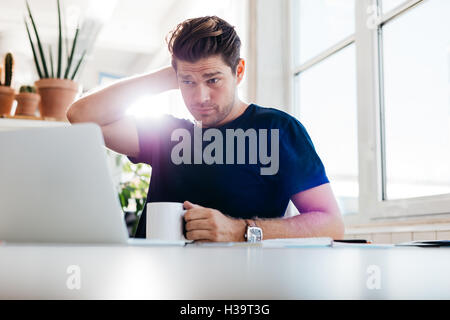 Junger Mann hält Kaffeetasse und Blick auf Laptop beim sitzt an seinem Schreibtisch im Büro. Durchdachten Business-Mann bei seiner Arbeit plac Stockfoto