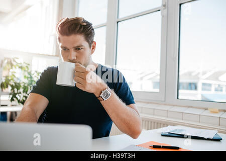 Junger Geschäftsmann, trinken Kaffee und arbeiten am Laptop im Büro. Männliche Führungskraft arbeitet an seinem Schreibtisch und Kaffee trinken. Stockfoto