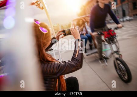 Frau fotografieren Freunde fahren Dreirad unterwegs. Junge Menschen auf Dreirad auf Stadtstraße. Stockfoto