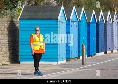 Weibliche Rasse Marschall in hohen Vis Weste stehend von Schattierungen von blau Strandhütten an Promenade, Bournemouth im Oktober Stockfoto