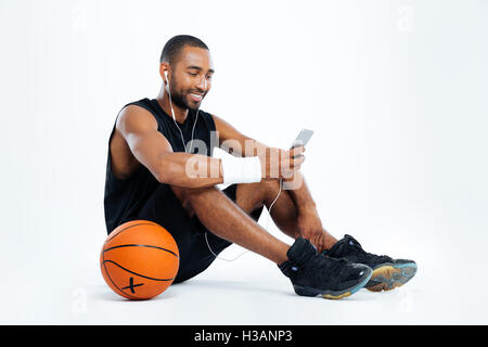 Fröhlicher junge Mensch-Basketball-Spieler sitzen und hören Musik vom Mobiltelefon auf weißem Hintergrund Stockfoto