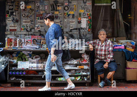 Frau vorbei ein Greis, der Zeitungen und Zeitschriften in Sheung Wan auf Hong Kong Island in Hongkong, China verkauft. Stockfoto
