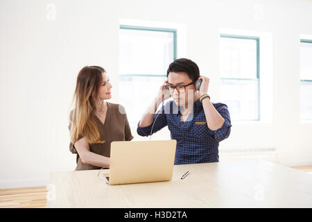 Geschäftsfrau, die männlichen Kollegen ansieht, die Kopfhörer tragen, während sie im Büro einen Laptop benutzt Stockfoto