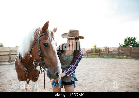 Fröhliche, nette junge Frau Cowgirl stehend und kümmert sich um ihr Pferd im Dorf Stockfoto
