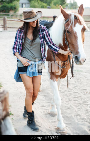 Fröhliche, nette junge Frau Cowgirl zu Fuß mit ihrem Pferd im Dorf Stockfoto