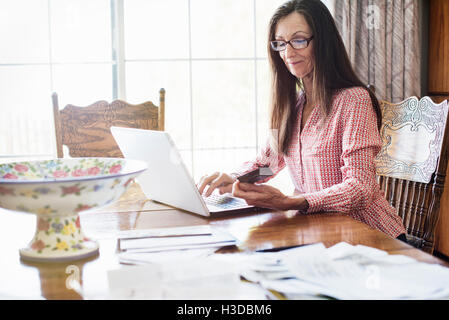 Senior Frau mit langen braunen Haaren sitzen an einem Tisch mit einem Laptop-Computer, Lesebrille tragen. Stockfoto