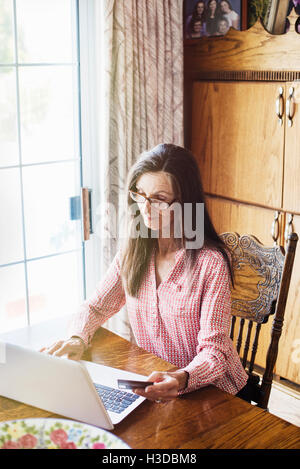 Senior Frau mit langen braunen Haaren sitzen an einem Tisch mit einem Laptop-Computer, Lesebrille tragen. Stockfoto