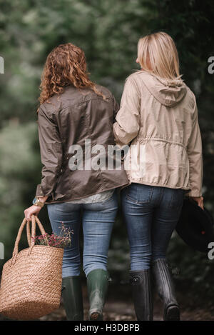 Zwei Frauen arm in Arm gehen in Mänteln und Stiefeln entlang einem Feldweg mit einem Korb. Stockfoto