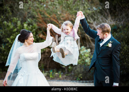 Eine Braut und Bräutigam, ein paar am Tag ihrer Hochzeit halten und schwingen ein kleines Mädchen in der Luft zwischen ihnen. Stockfoto