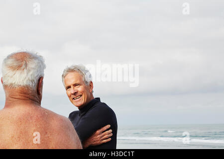 Zwei ältere Männer stehen an einem Strand im Chat, einen Neoprenanzug tragen. Stockfoto