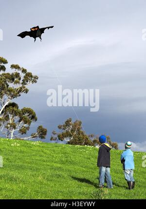 Zwei jungen fliegen eine schwarze Fledermaus Kite am Fuße eines grünen Hügels, mit dunklen grauen Wolken im Hintergrund. Stockfoto