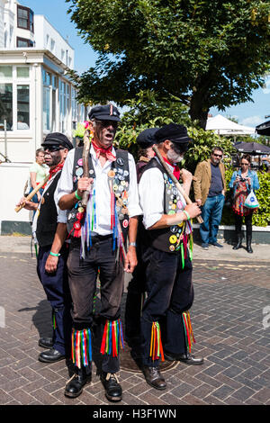 Traditionelle Volkstänzer, das tote Pferd Morris Men mit geschwärzten Gesichtern tanzen im Freien in der Sonne, während Verantwortung Holzpfähle. Stockfoto