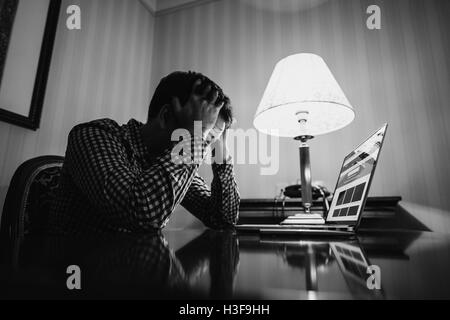 Erschöpft. Frustrierter junge Mann mit Händen auf dem Kopf und Augen zu halten beim Sitzen am Arbeitsplatz geschlossen. Schwarz weiß Foto. Stockfoto