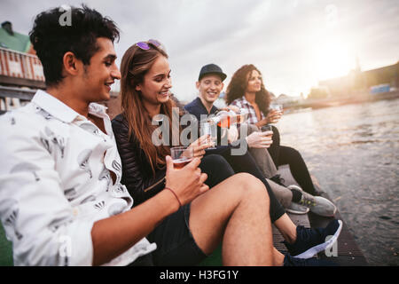 Schuss Gruppe von Menschen sitzen im Freien an einem Anlegesteg und Getränke. Freunde auf Pier über See in der Stadt hängen. Stockfoto