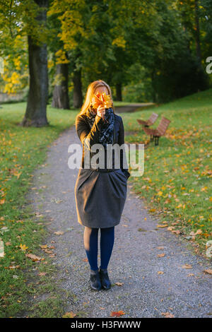 Junge Dame stehend auf Pfad im herbstlichen Park und Gesicht hinter Haufen Laub versteckt Stockfoto